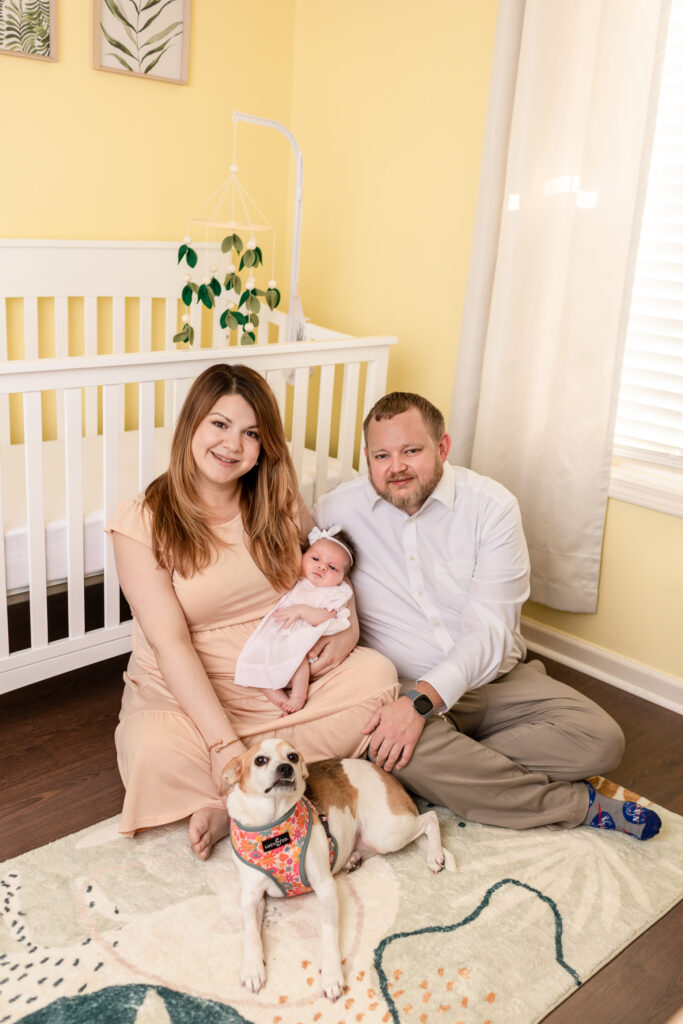 Parents with baby girl and dog sitting on the floor in the nursery during their in-home newborn photography session.