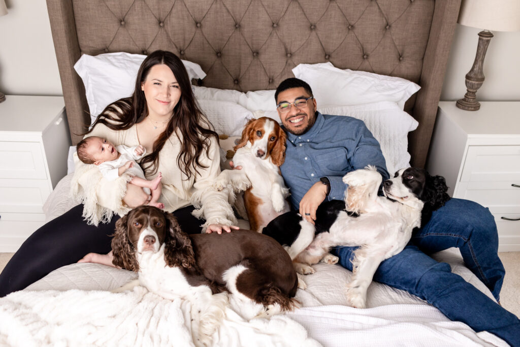 New parents sitting on bed with newborn baby girl and dogs during their in-home newborn photography session.