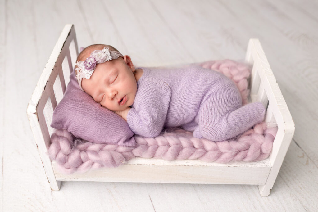 Baby girl in a knit footed sleeper posed on a tiny bed during her in-home newborn photography session in Frankfort, Kentucky.