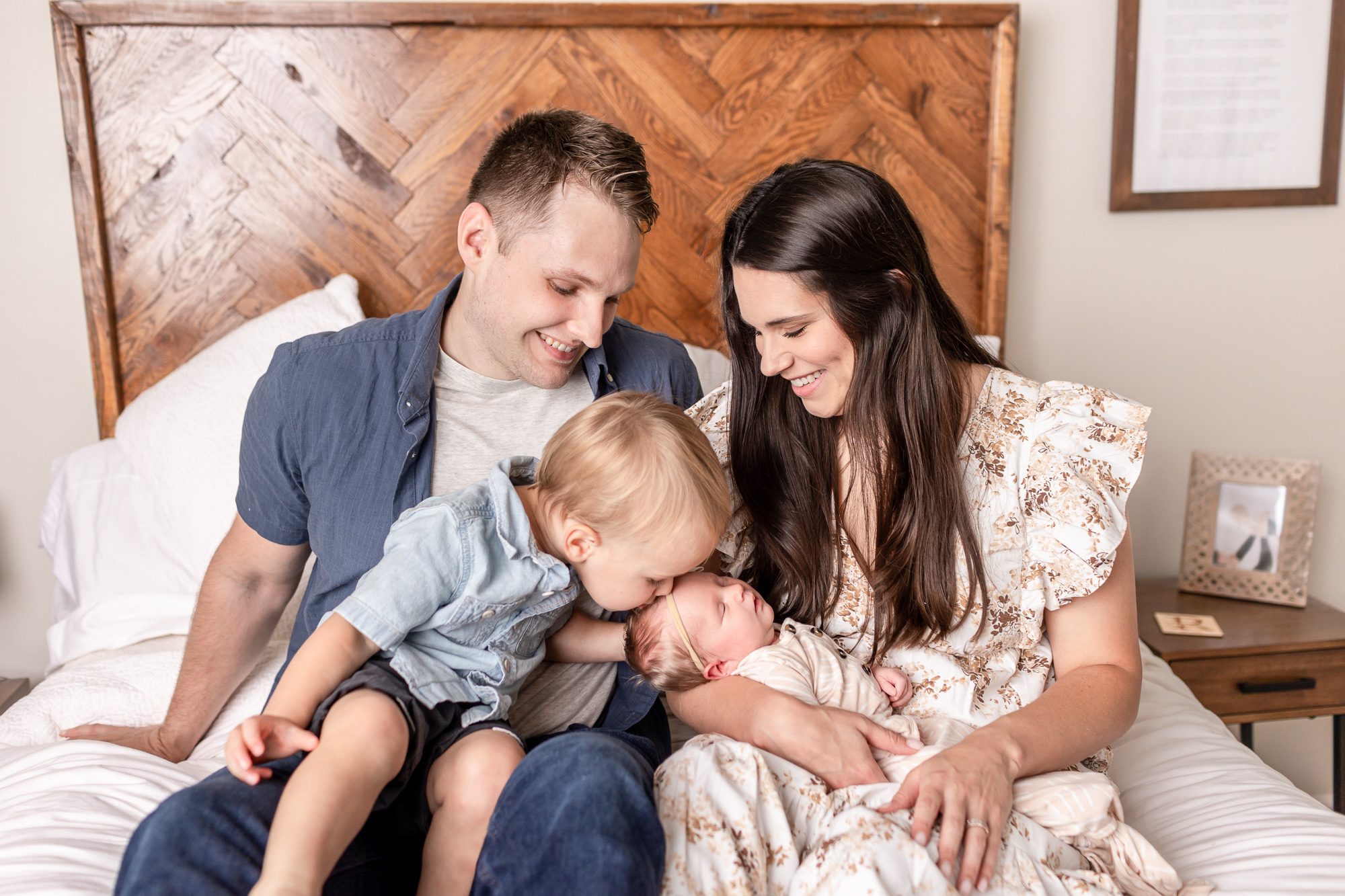 Family sitting on bed during their in-home newborn photography session. Big brother is kissing baby sister.